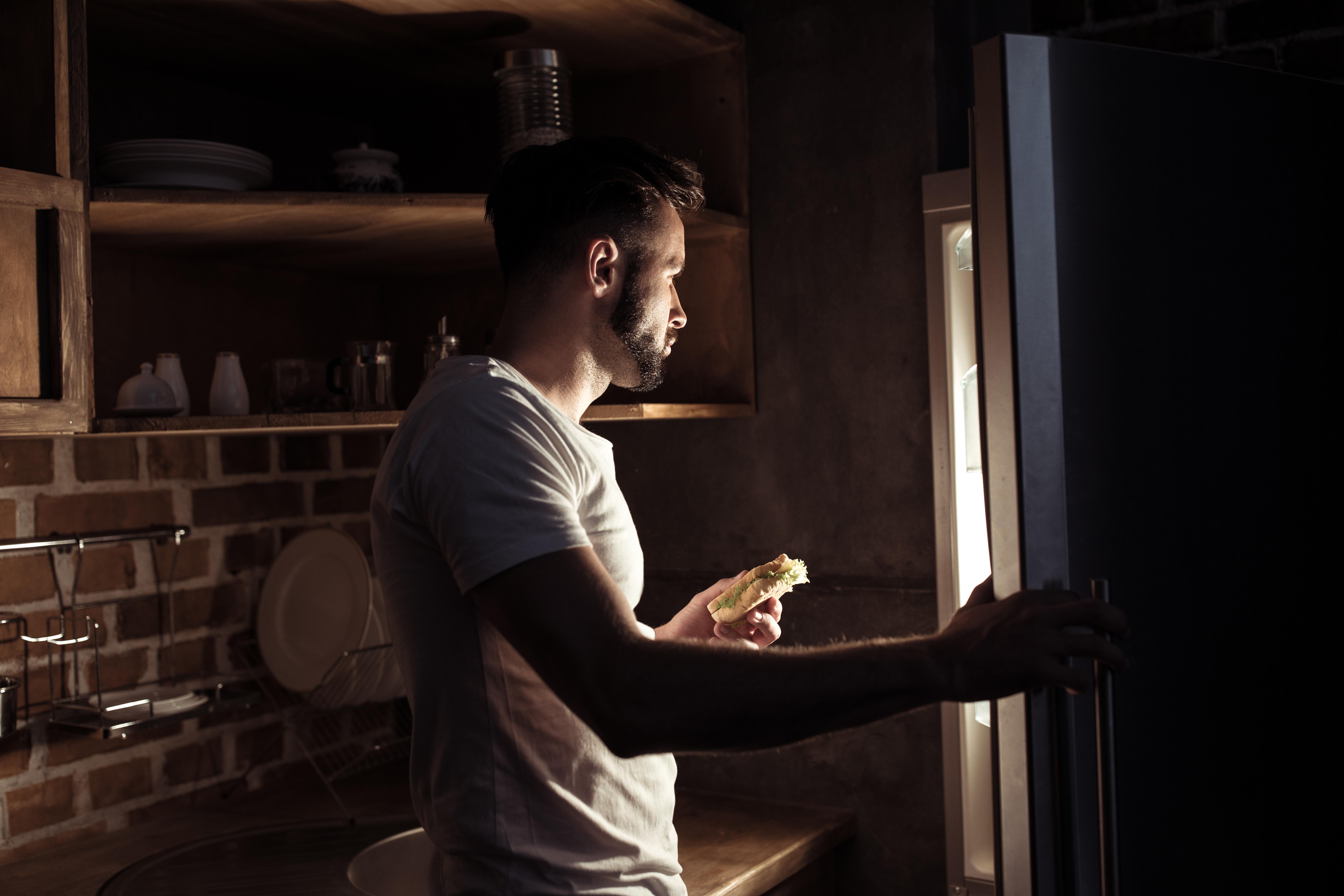 man looking into refrigerator holding sandwich