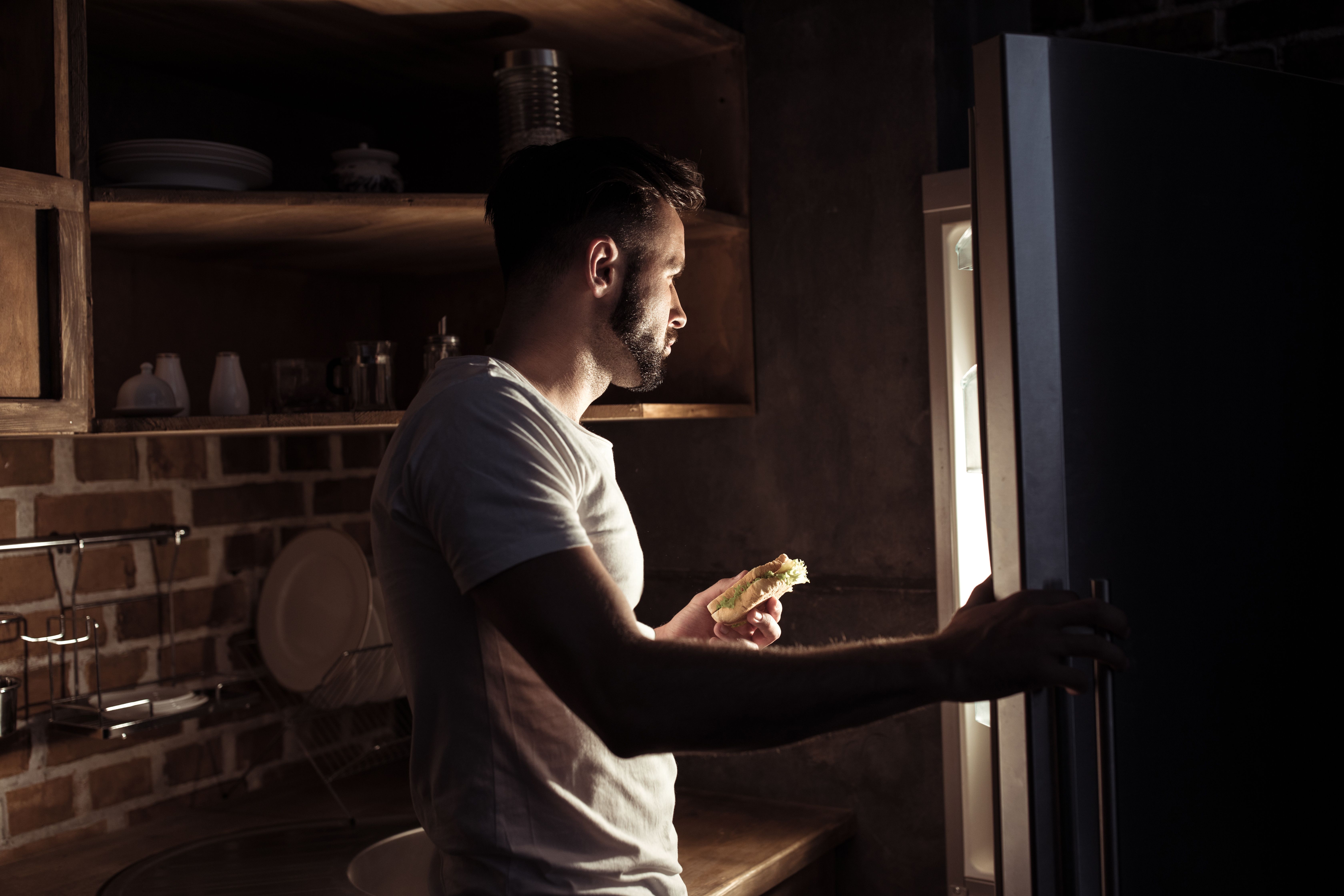 man looking into refrigerator holding sandwich