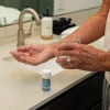 older man holding a pill in one hand and a glass of water in the other by the sink in a bathroom with capsules packaging by the sink